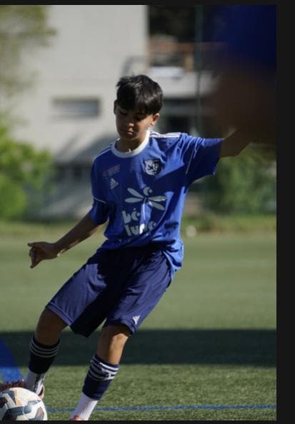 Jeune garçon en tenue de football bleue dribblant un ballon sur un terrain ensoleillé.