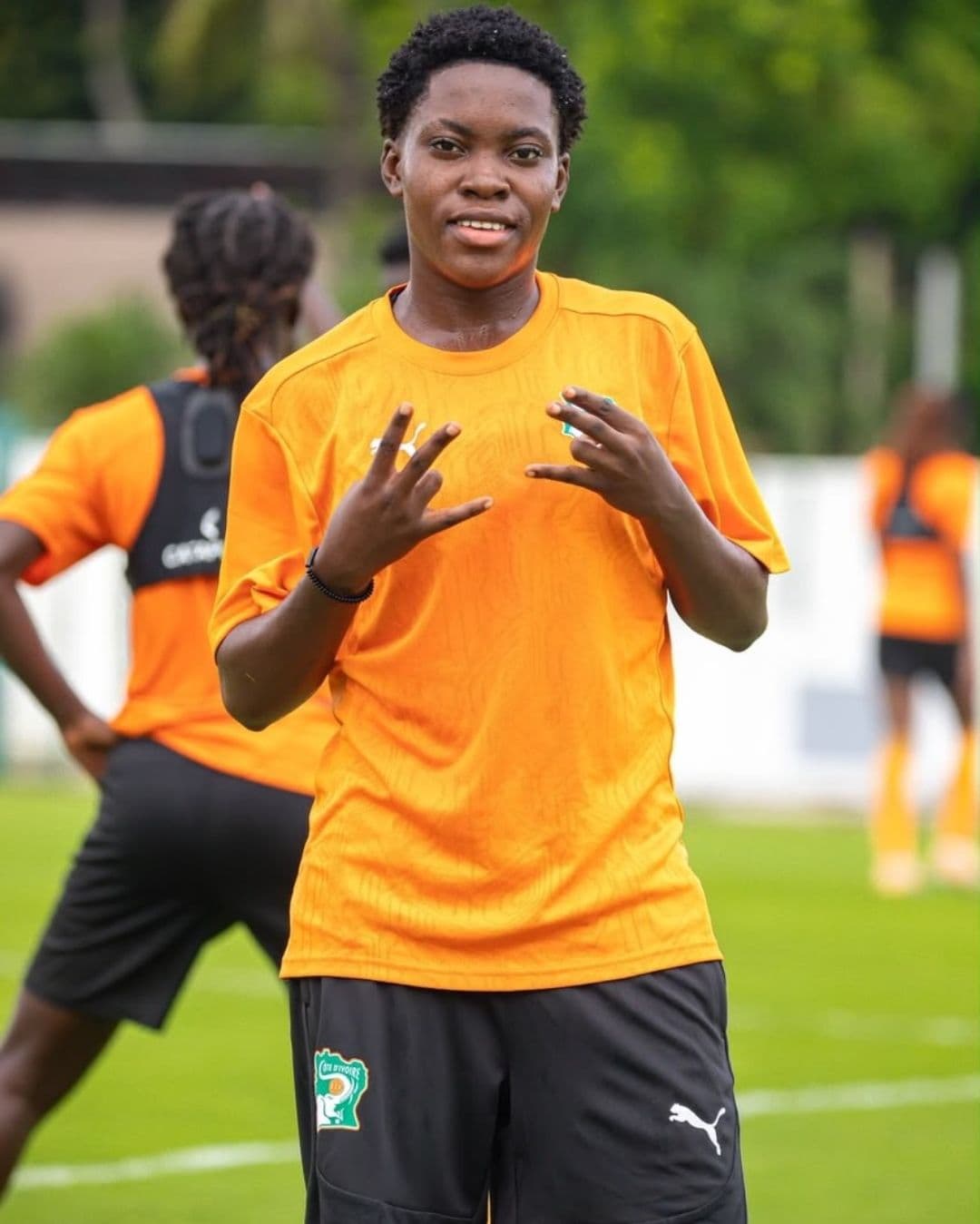 Footballeuse ivoirienne en maillot orange faisant des signes de la main sur un terrain.