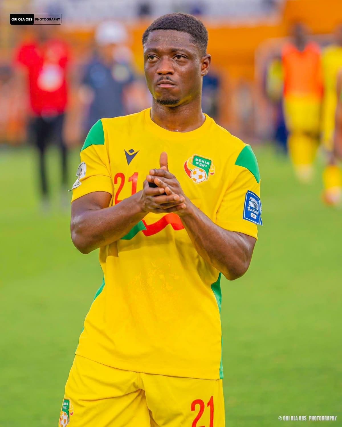 Footballeur béninois en maillot jaune numéro 21, mains jointes sur un terrain de football.