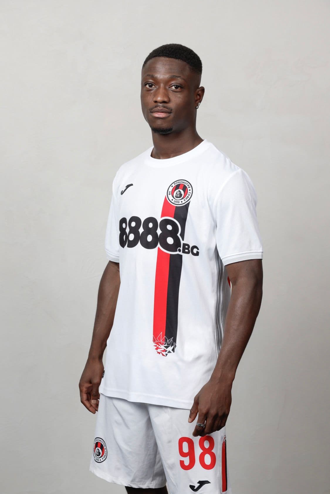 Footballeur noir en maillot blanc avec bande verticale rouge et noire, posant sur fond gris.