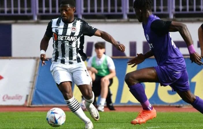 Footballeur en maillot rayé noir et blanc dribblant le ballon devant un adversaire en violet.
