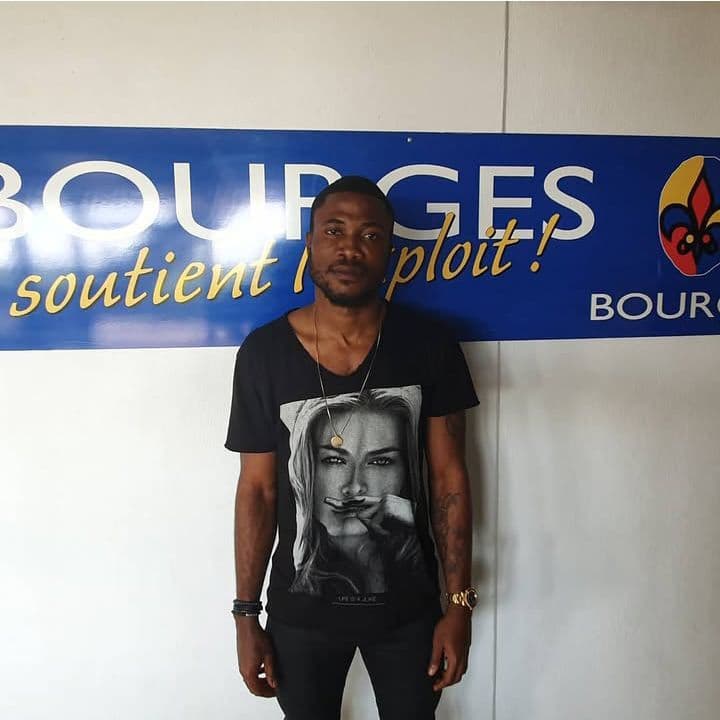 Homme en t-shirt noir avec portrait, posant devant une affiche bleue "Bourges soutient l'exploit !"