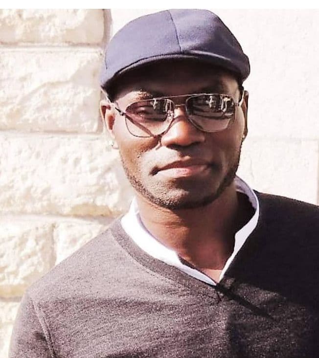 Homme noir avec casquette plate et lunettes de soleil devant un mur en pierre.