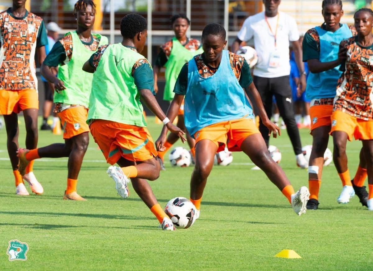 Des joueuses de football s'entraînent avec un ballon sur un terrain de sport ensoleillé.