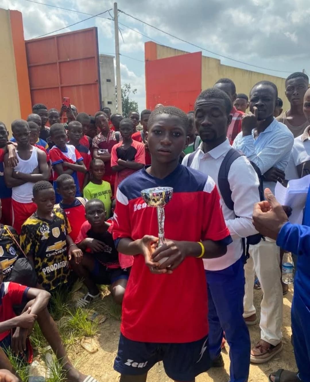 Jeune garçon en maillot rouge tenant un trophée argenté devant une foule de spectateurs.